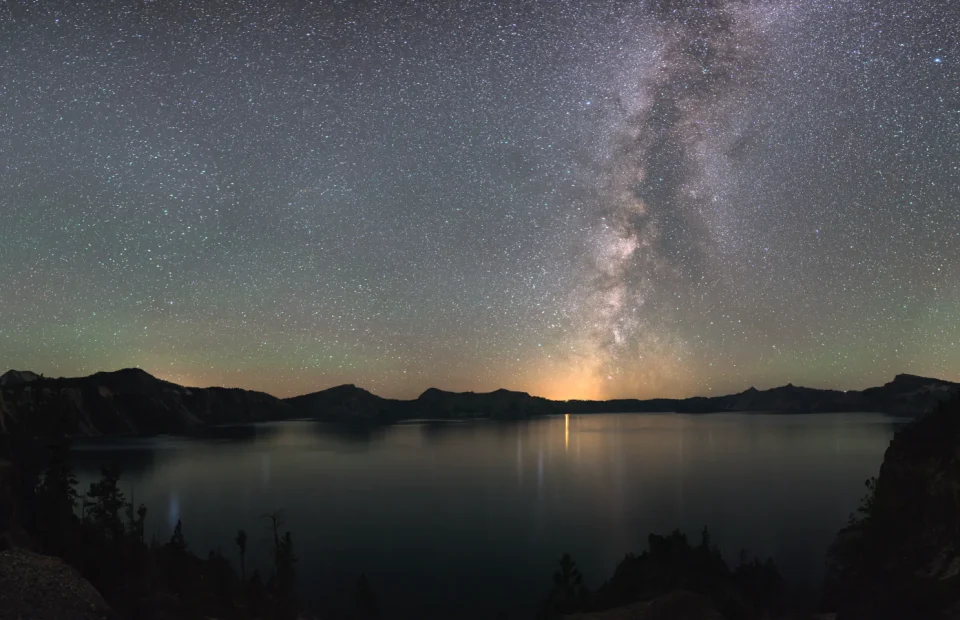The Milky Way rises above the dark caldera and still water of Crater Lake National Park